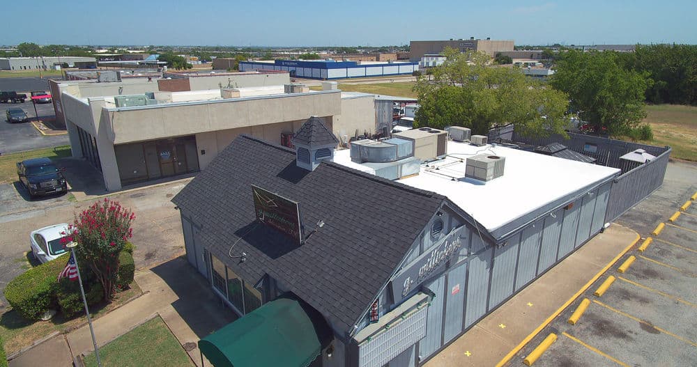 Aerial view of a commercial building with a flat roof, surrounded by parking and nearby structures.
